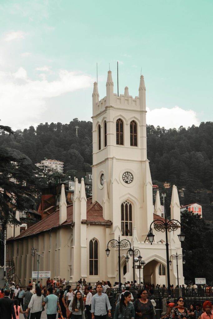 The Christ Church in Shimla, a popular winter tourist destination in India, surrounded by lush green hills and a crowd of visitors. The iconic neo-Gothic architecture stands tall against the serene sky, with the area bustling with activity, making it a perfect spot for travelers exploring India's winter retreats.