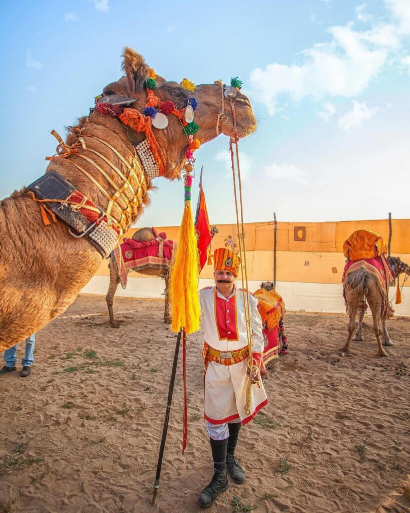 A cameleer standing beside a camel in the golden sands of the Jaisalmer desert, offering rides to tourists. The vast desert landscape and traditional attire of the cameleer highlight the cultural charm of Jaisalmer, a popular winter tourist destination in India known for its desert safaris and rich heritage.