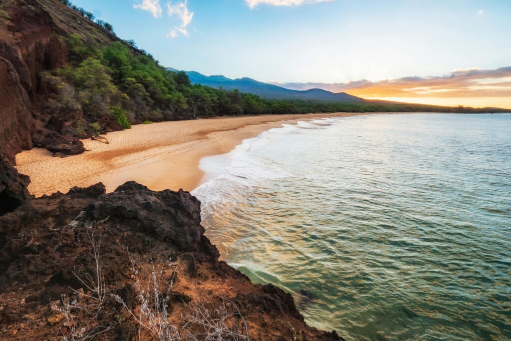 Stunning Maui beach at sunset, with golden and pink hues reflecting off the calm ocean waters and casting a warm glow on the sandy shore, creating a romantic setting ideal for anniversary trip ideas.